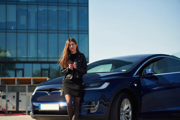 In black jacket and skirt. Young woman is near her electric car outdoors