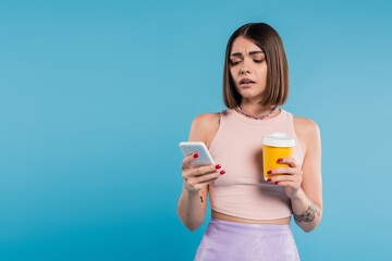 to go coffee, social media influencers, tattooed young woman with short hair and nose piercing holding paper cup and using smartphone on blue background, generation z, summer trends