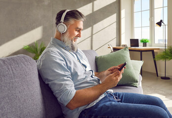 Side view portrait of handsome bearded mature man in headphones listening to favourite music using his smartphone, sitting on the sofa having rest and relaxing in the living room at home.