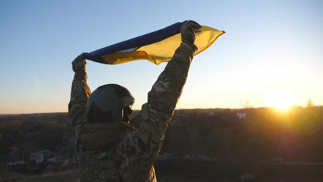 Dolly shot of female soldier of ukrainian army a raised over head blue-yellow banner at sunset. Young girl in camouflage uniform lifted flag of Ukraine at countryside. Invasion resistance concept