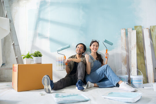 Portrait Asian couple repairing and painting the wall with blue paint using a roller during renovation in their new apartment.