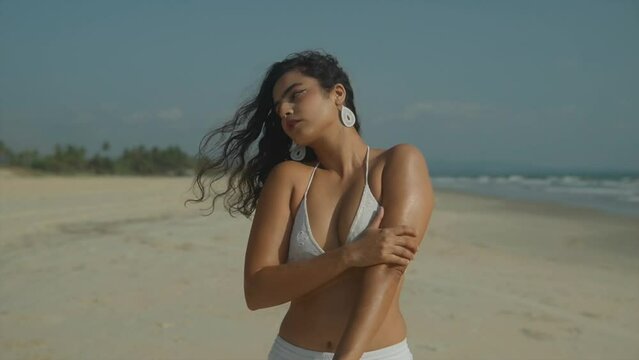 South Asian woman wearing white bikini applying suncream to arms while on sunny beach