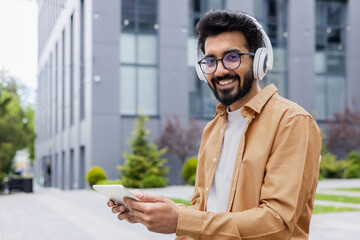 Portrait young Indian man with headphones and tablet watching online video sitting on bench near office building businessman smiling and looking at camera, portrait of satisfied online stream viewer - Powered by Adobe
