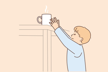 Boy reaches for mug of hot tea on table at risk of getting burned and getting into trouble
