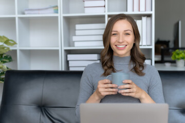 Cute young woman using laptop computer sitting on sofa in the living room and holding coffee, watching movie, listening to music, shopping online, watching live broadcast.