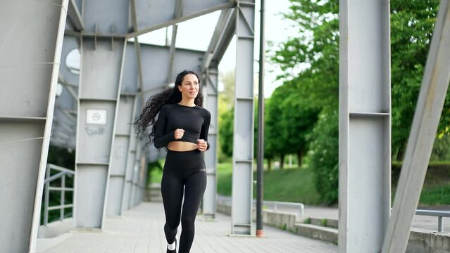 Young adult female runner jogging on the street in the city. Happy brunette sportswoman enjoys jogging outdoors. A fit woman in a tracksuit is doing her daily jog outdoors running on an urban stadium