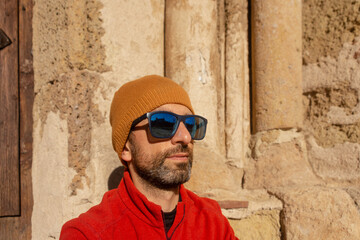 Close-up of the face of a man with a beard wearing a beanie and sunglasses, with neutral expression an old stone wall in the background
