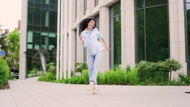 Happy joyful young and attractive woman dancing walking on the street in front of a modern building. Active pretty curly brunette female cheerfully celebrating achievement. Dance of a funny teenager