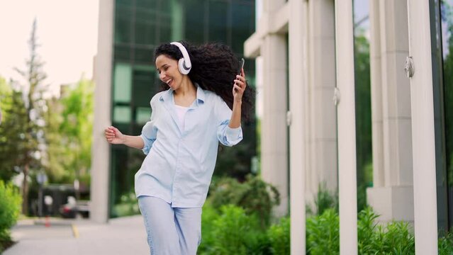 Happy joyful young attractive woman dancing walking on the street in front of a modern building. Active pretty curly brunette female enjoys listening to music on headphones. Dance of a funny teenager