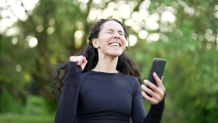 Happy excited young sportswoman checking the results of training while looking at smartphone in urban city park. Smiling female happy with good successful workout results or great news on mobile phone