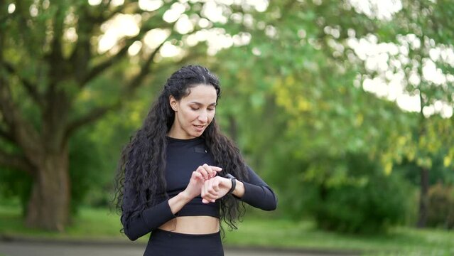 Excited smiling young woman runner looking smart watch in urban city park. Sportswoman using checking fitness tracker. Fit female is satisfied with the results of training, rejoices, shows yes gesture