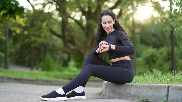 Smiling young sportswoman looking smart watch sitting in urban city park. Woman using checking fitness tracker. Fit female is satisfied with the results of training, rejoices, shows a yes gesture