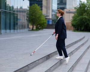 Blind business woman in glasses and with a cane climbs the stairs to the business center. 