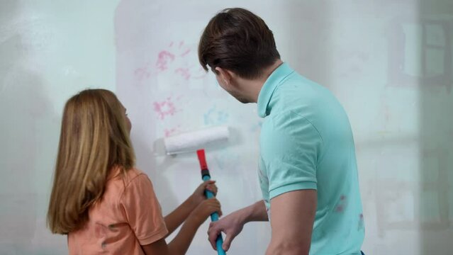 Joyful father touching nose of smiling daughter helping painting wall in living room. Cheerful Caucasian young man and adolescent girl having fun repairing house indoors