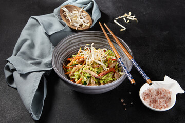 An appealing chicken salad with vegetables, Peking cabbage, and soy sprouts in an Asian style bowl, served with chopsticks on a black background. Accented with pink sea salt