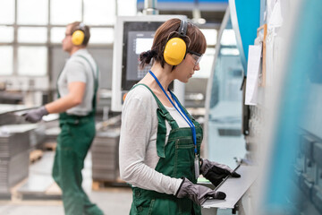 Female worker with ear protectors holding metal part in factory - obrazy, fototapety, plakaty