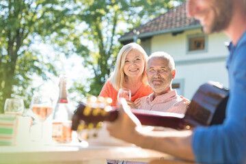 Senior couple drinking wine watching adult son playing guitar on sunny patio