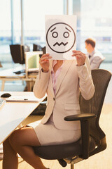 Portrait businesswoman holding frowning face printout over her face in office