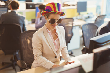 Playful businesswoman wearing silly sunglasses striped hat at computer in office