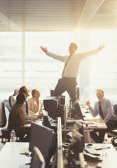 Colleagues watching exuberant businessman celebrating on top of desks in office