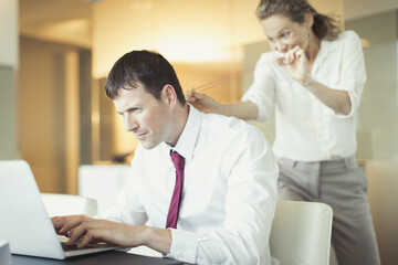 Mischievous businesswoman aiming rubber bat unsuspecting businessman working at laptop
