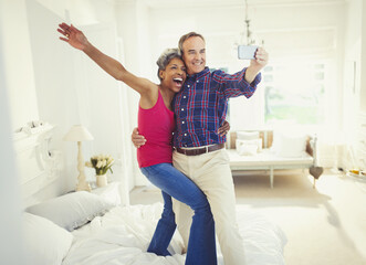 Playful mature couple taking selfie standing on bed