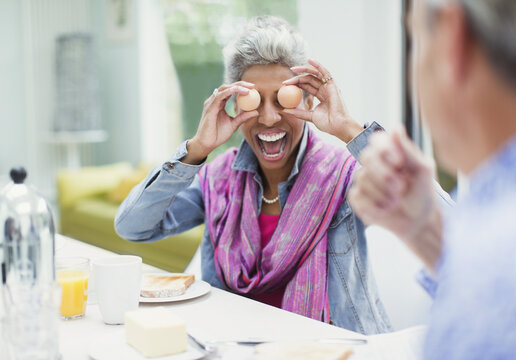 Playful mature woman covering eyes with eggs at breakfast table