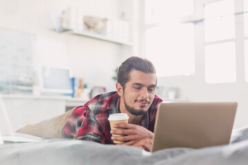 Young man drinking coffee at laptop on bed - obrazy, fototapety, plakaty