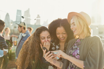 Young women laughing and texting with cell phone at rooftop party