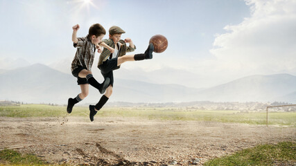 Active, cheerful, little boys, children in retro style clothes kicking ball in jump, playing football outdoors on a daytime