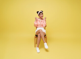 Attractive beautiful Asian woman sitting on chair and hands up raised arms from happiness isolated on yellow background. Excited student woman winner success concept.
