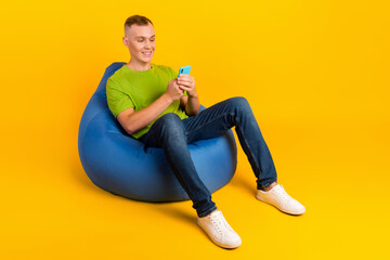 Cheerful attractive guy holding smart phone sitting bean bag pouf using 3G internet over yellow...