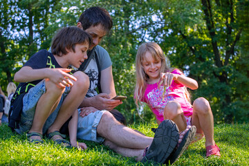happy father with son and daughter look at the gadget information while sitting on the grass in nature. Joint family vacation. Two generations father and son and daughter