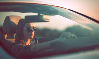 Young woman wearing glasses behind the wheel of a convertible during sunset.