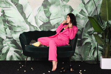 Young woman in pink suit spending time alone at home eating popcorn on the couch looking tv. messy...