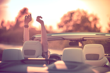 Relaxing in a convertible during sunset. Happy young woman with her hands up.