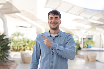 young handsome man smiling cheerfully, feeling happy and pointing to the side and upwards, showing object in copy space