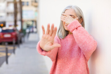 senior retired pretty white hair woman covering face with hand and putting other hand up front to...
