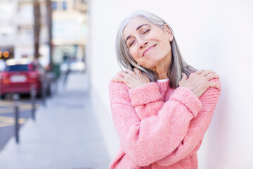 senior retired pretty white hair woman feeling in love, smiling, cuddling and hugging self, staying...