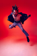 Full-length portrait of young punk in extraordinary clothes, hairstyle and makeup posing over red studio background in neon light. Concept of music, lifestyle, subculture, art, youth, human emotions