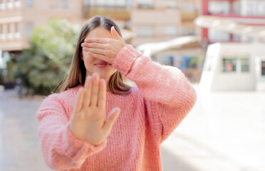 young pretty woman covering face with hand and putting other hand up front to stop camera, refusing...