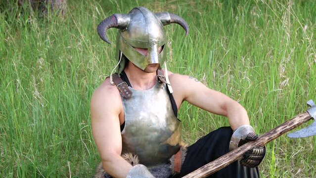 Smooth camera movement, selective focus. Portrait of a warrior in a horned helmet, a steel breastplate with a two-handed ax in his hands, sitting in tall grass against the backdrop of the forest.