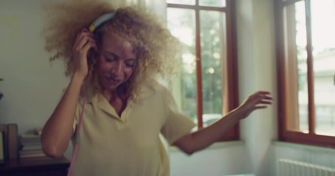 Young Stylish Brazilian Woman With Headphones on and Dancing to the Beat of a Song in her Apartment. Beautiful Carefree Female with Curly Hair Enjoying her Time By Moving Along Happy Music