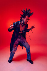 Full-length portrait of young punk in extraordinary clothes, hairstyle and makeup posing over red studio background in neon light. Concept of music, lifestyle, subculture, art, youth, human emotions