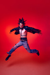 Fototapeta na wymiar Full-length portrait of young punk in extraordinary clothes, hairstyle and makeup jumping over red studio background in neon light. Concept of music, lifestyle, subculture, art, youth, human emotions