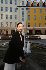 Beautiful young brunette in white dress and black blazer walking outdoors on the street of an European city against a fountain and fancy old buildings
