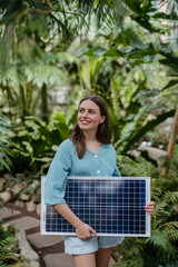 Young woman in jungle holding paper model of house with solar panels, concept of renewable energy and protection of nature.