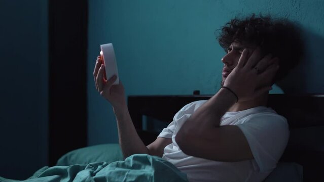 Frustrated young man in bed suffering insomnia, looking on alarm clock and holding his head