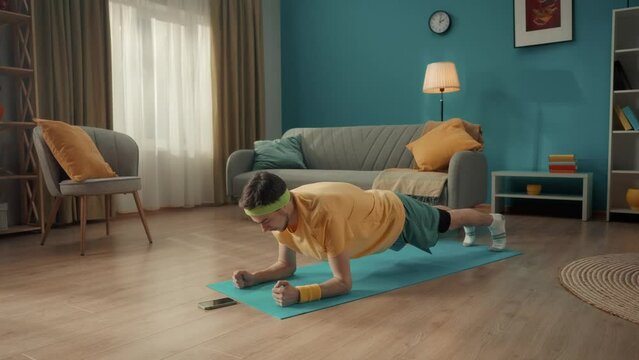 A man in sportswear is lying on the floor in front of the phone. A man starts a timer on his smartphone and gets into a plank position on his elbows. Home fitness concept, online sports class.