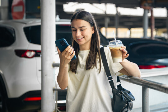 Cheerful young woman with smartphone and something in the parking lot.